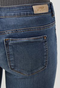 Temno modre jeans hlače iz denima z teksturiranim zaključkom, z vidnimi šivi, z oznako na nalepki nad zadnjim žepom in klasičnim dizajnom s petimi žepi.