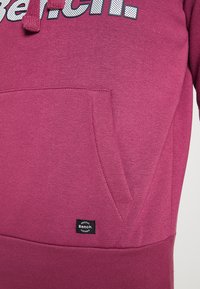 Bordeauxroter Hoodie mit einer vorderen Kängurutasche, ausgestattet mit einem kontrastierenden weißen Logo. Weiche Textur mit geripptem Saum und Etikett-Detail.