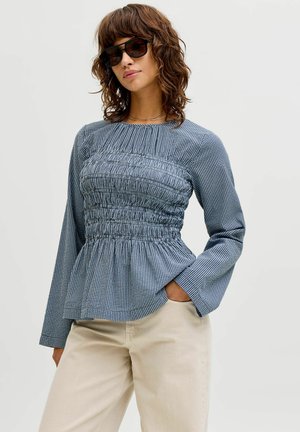 Femeie purtând ochelari de soare negri supradimensionați, o bluză albastră cu mâneci lungi și pantaloni bej, pozând cu o mână în buzunar pe un fundal simplu.