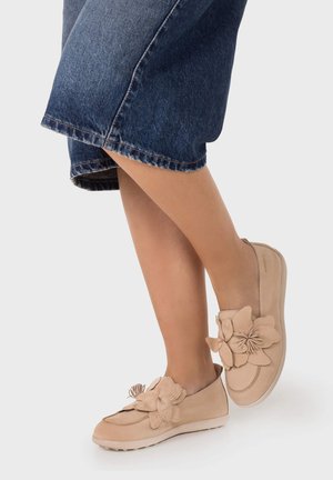 Personne portant des chaussures beige sans lacets avec de grandes décorations florales, associées à un pantalon en denim bleu foncé ample, sur un fond uni.