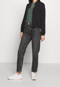 Giubbotto bomber nero con polsini a coste, blusa verde, jeans scuri a vita alta e sneakers bianche. Texture lisce e design aderente.