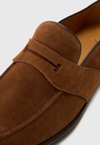 Mocassins en daim marron avec une bride selle, une couture lisse et un bout arrondi. L'intérieur est en cuir marron clair avec une semelle polie.