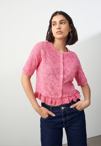 Kurzärmelige rosa Spitzenstrickjacke mit Blumenmuster, welligem Saum und Knopfverschluss. Kombiniert mit dunkelblauer Jeans, Hände in den Taschen.