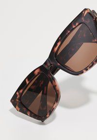 Óculos de sol de padrão tartaruga com uma armação geométrica, lentes com tonalidade castanha e bordas grossas; padrões texturizados visíveis nas laterais da armação.