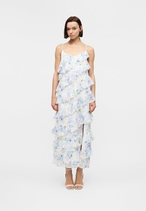 Kobieta stojąca w długiej białej sukience w kwiaty z warstwowymi falbanami i cienkimi ramiączkami, w połączeniu z beżowymi sandałami na wysokim obcasie.