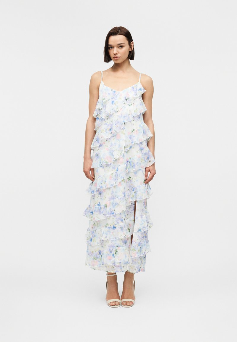 Γυναίκα στέκεται φορώντας λευκό μακρύ φόρεμα με λουλουδένιο μοτίβο, στρώσεις βολάν και λεπτές τιράντες, συνδυασμένο με μπεζ πέδιλα με ψηλό τακούνι.