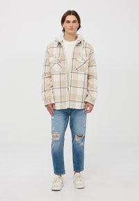 Giacca a quadri beige con due tasche sul petto e cappuccio grigio, abbinata a jeans strappati blu chiaro e sneaker bianche.