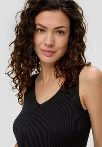 Kobieta z kręconymi ciemnymi włosami, ubrana w czarny bezrękawnik z prążkowanego materiału, patrzy bezpośrednio w aparat z lekkim uśmiechem.