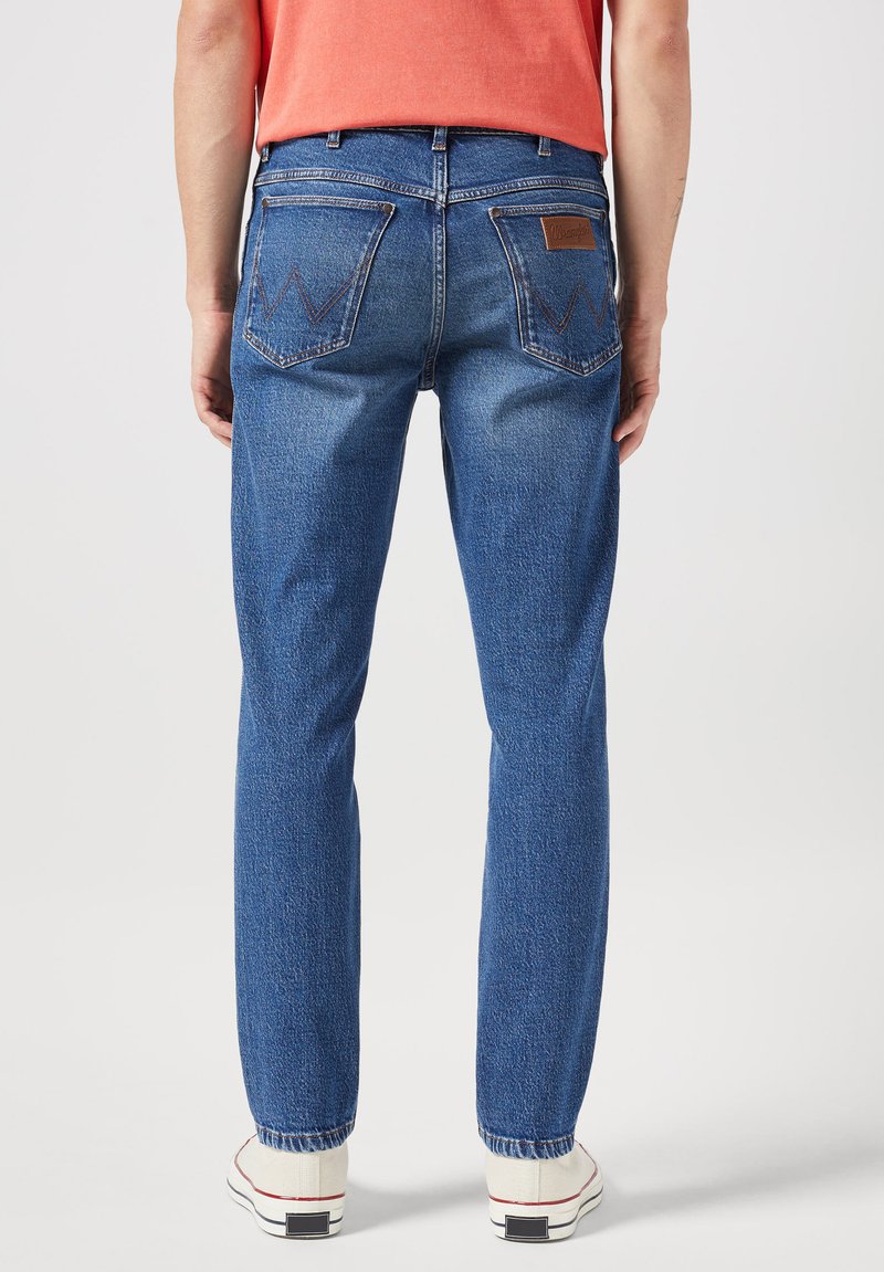 Vaqueros de mezclilla azul con corte recto, que cuentan con dos bolsillos traseros con costura en zigzag y una etiqueta de cuero marrón en la cintura.