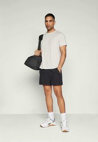 Lys grå t-skjorte, svarte shorts, hvite joggesko med svarte detaljer. Svart treningsbag hengende over den ene skulderen. Minimalistisk design, avslappet passform.