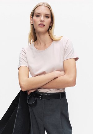 Kvinde med blondt hår iført lysrosa kortærmet skjorte, grå bukser med sort bælte, holder en sort jakke over armen, foran hvid baggrund.