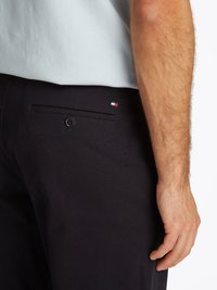 Calças de marinha com um bolso traseiro que apresenta um botão preto e um pequeno logotipo em vermelho, branco e azul. O tecido parece suave e resistente.