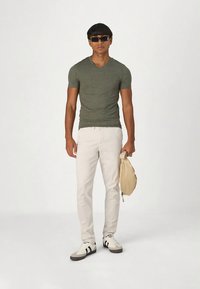 Olivgrünes V-Ausschnitt-T-Shirt, hellbeige Hose und weiße Sneaker mit schwarzen Streifen; hält eine beige Tasche; steht vor einem schlichten Hintergrund.