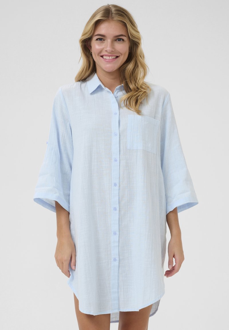 Robe chemise bleu clair en tissu texturé. Elle possède un col, une poche avant et des manches courtes amples.
