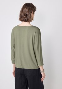 Maglietta verde oliva a maniche lunghe con una vestibilità comoda, caratterizzata da polsini arrotolati e scollatura arrotondata, realizzata in tessuto morbido e leggero.