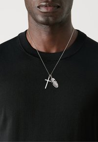 Sølv halskjede med kors og fjær charms, båret over en svart teksturert genser med rund hals. Høy kontrast mellom materialene.
