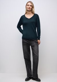 Maglione verde scuro a scollo a V con maniche lunghe, realizzato in un tessuto morbido, abbinato a jeans grigi a gamba dritta e stivaletti neri alla caviglia.