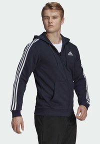 Sudadera con cremallera de color azul marino con rayas blancas de Adidas en las mangas, bolsillos delanteros y capucha con cordón. Fabricada en material suave y texturizado.