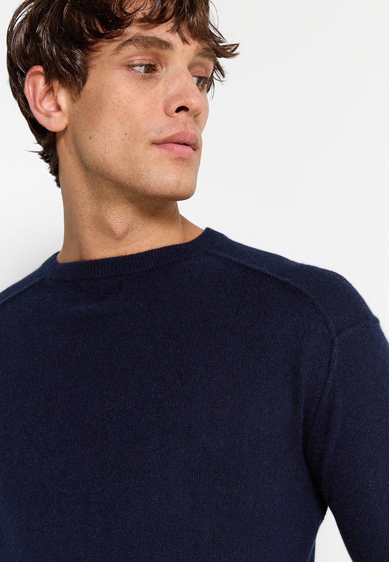 Granatowy sweter z okrągłym dekoltem wykonany z miękkiego dzianiny, z teksturowanym wykończeniem i ściągaczami na mankietach, noszony przez mężczyznę.