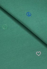 Tessuto verde con piccoli simboli ricamati: una faccina sorridente verde chiaro, un simbolo della pace blu e un cuore rosa.