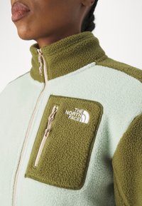 Fleecejacka med mintgrön kropp och mörkgröna detaljer, hög krage och en dragkedjeförsedd bröstficka med logotyp. Strukturerad tyg.