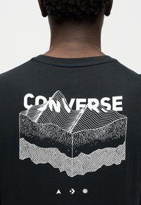 Μαύρο βαμβακερό μπλουζάκι με λευκό σχέδιο στρωμένων βουνών και τη λέξη "CONVERSE" με έντονα γράμματα πάνω, με τριγωνικά, αστεροειδή και σφαιρικά εικονίδια κάτω.