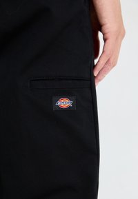 Black pants made of textured fabric, featuring a side pocket and a rectangular embroidered Dickies logo patch on the upper leg.