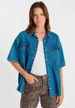 Femme portant une chemise en jean ample à manches courtes par-dessus un débardeur blanc avec un pantalon à imprimé léopard, debout avec une main dans la poche.