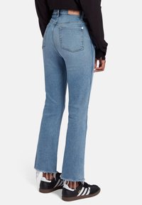 Jeans a zampa d'elefante blu chiaro con orlo sfilacciato, cinque tasche e lavaggio sbiadito, abbinati a sneaker nere con strisce bianche.