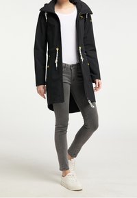 Black waterproof coat with a zip front, front pockets, adjustable drawstrings, and gold-tone hardware, paired with gray jeans and white sneakers.