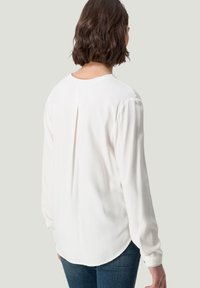 Femme aux cheveux courts bruns vue de dos, portant un chemisier blanc à manches longues et un jean bleu foncé sur un fond clair.
