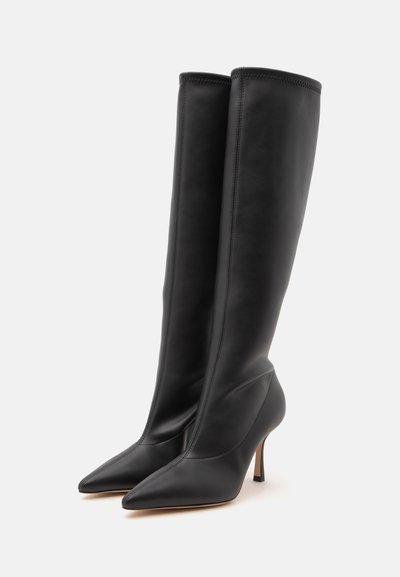Paire de bottes en cuir noires montantes jusqu’au genou avec des bouts pointus et des talons hauts et fins sur un fond clair uni.
