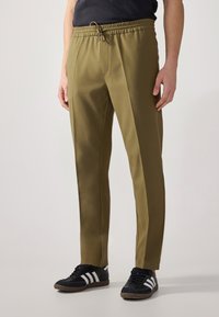 Pantaloni verde oliva con vita elastica, dettaglio con coulisse e strisce laterali. Abbinati a scarpe da ginnastica nere con accenti bianchi.