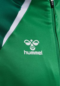 Groene sportjacket met een gladde textuur, witte accenten en een prominent wit 'hummel' logo met een gestileerd insectontwerp.
