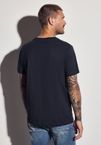 Muž s tetovanými rukama oblečený v černém tričku a modrých džínách, stojící a dívající se na stranu na bílém pozadí.