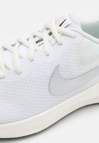 Białe sportowe sneakersy z siatkowego materiału, białymi sznurówkami i szarym logo Nike w kształcie „swoosh” po boku.