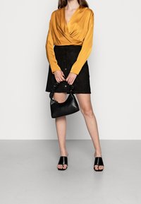 Satynowy top w kolorze musztardowym z kopertowym dekoltem, mini spódniczka z czarnego zamszu z guzikami, czarna skórzana torebka z falbankowatym uchwytem oraz czarne klapki.