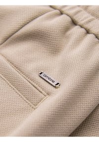 Beige textured fabric with a small silver metal tag reading "ombre" sewn near a stitched pocket edge.