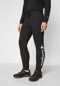 Sorte treningsbukser med slim fit, med snøring i livet og en hvit "Champion"-logo langs venstre ben i en krum skrifttype.