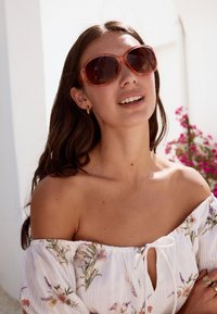 Oversized solglasögon med genomskinlig orange ram, mörka linser och blommig vit off-shoulder-topp med rosett.