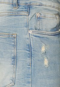 Acercamiento a un bolsillo trasero de unos vaqueros de mezclilla azul claro desgastados, con dos pequeños desgarros cerca del borde del bolsillo.