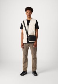 Beige Baumwollweste mit mehreren Taschen, getragen über einem schwarzen T-Shirt. Schwarze Umhängetasche mit Reißverschlüssen. Hellbraune Hose und schwarze Sneaker.