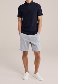 Navy polo shirt with three-button placket, paired with light gray patterned shorts. Model wearing white sneakers, standing in a neutral background.