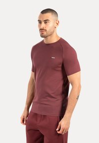 Man med kort hår och skägg, iklädd en åtsittande vinröd kortärmad skjorta och matchande byxor, stående framför en enkel ljus bakgrund.