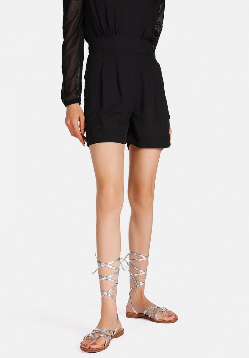 Pantaloni corti neri a vita alta con pieghe e una consistenza liscia, abbinati a sandali argentati con lacci che avvolgono le gambe inferiori.