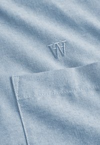 Light blue fabric with a subtle texture, featuring an embroidered "W" detail and a pocket outlined in a straight stitch.
