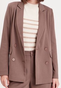 Blazer marron à double boutonnage en tissu doux, avec deux boutons, des revers pointus et des poches, porté sur un pull crème à rayures.