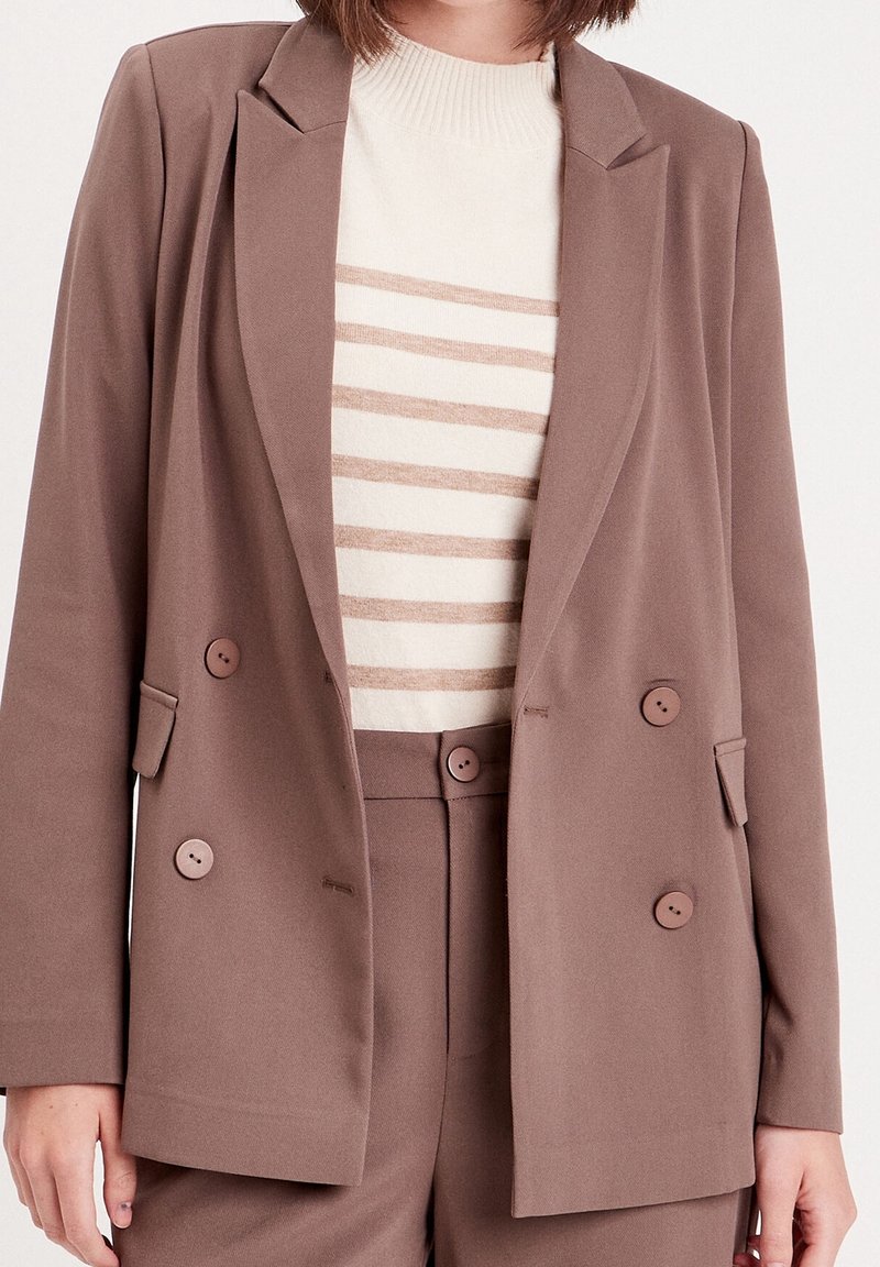 Blazer marron à double boutonnage en tissu doux, avec deux boutons, des revers pointus et des poches, porté sur un pull crème à rayures.
