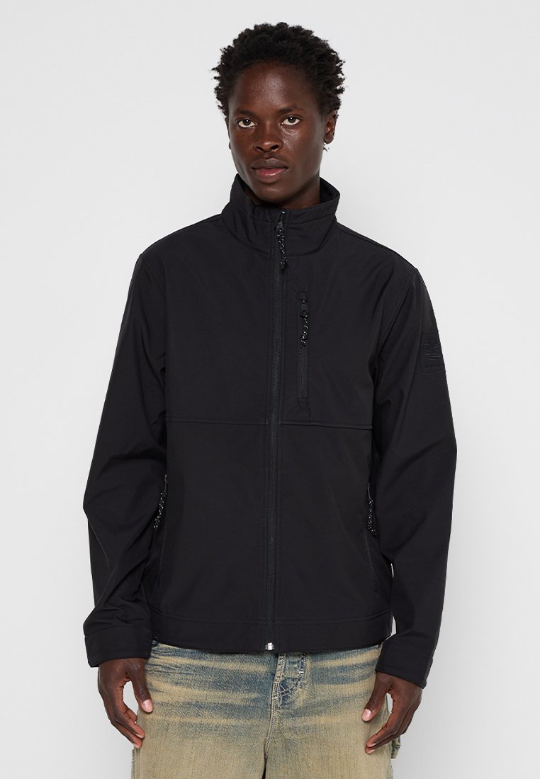 Man in een zwart ritsjack met hoge kraag en vervaagde blauwe jeans, staand tegen een effen lichte achtergrond.