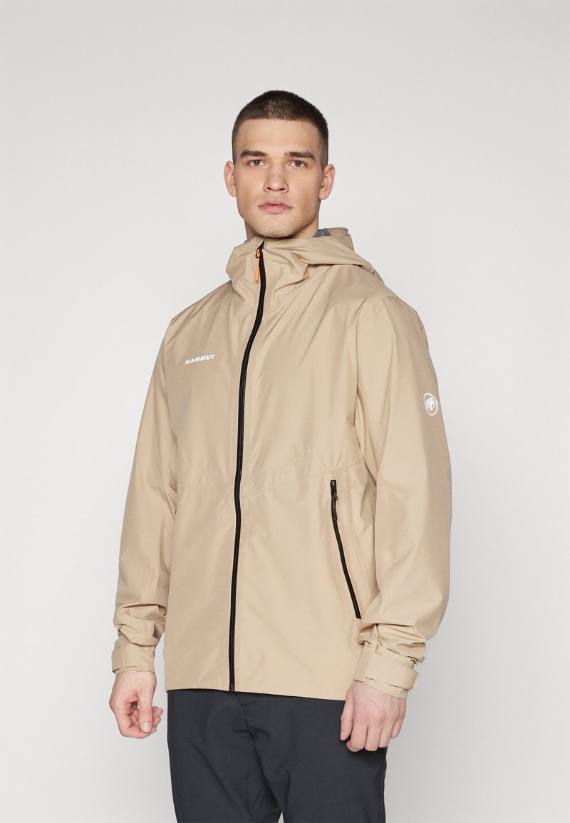 Beige vattentät jacka med frontdragkedja, justerbar huva och logotyp på bröstet. Har sidofickor och elastiska muddar.